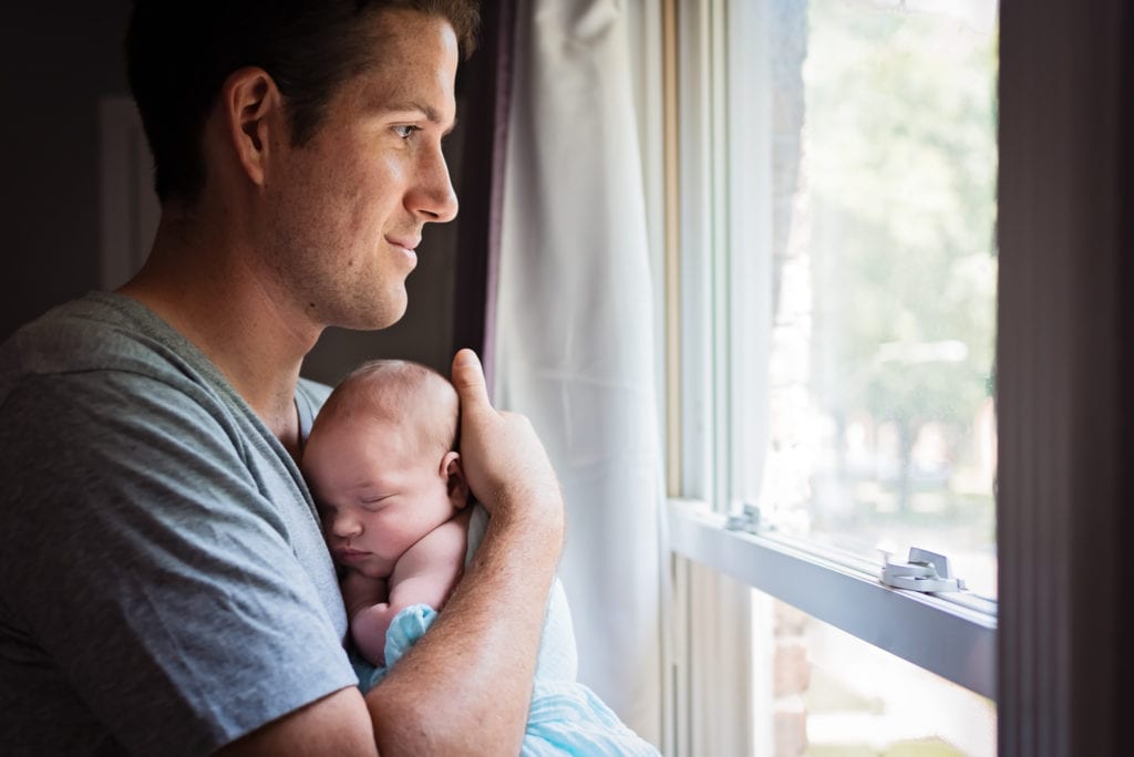 dad standing by window in grey t-shirt holding newborn baby