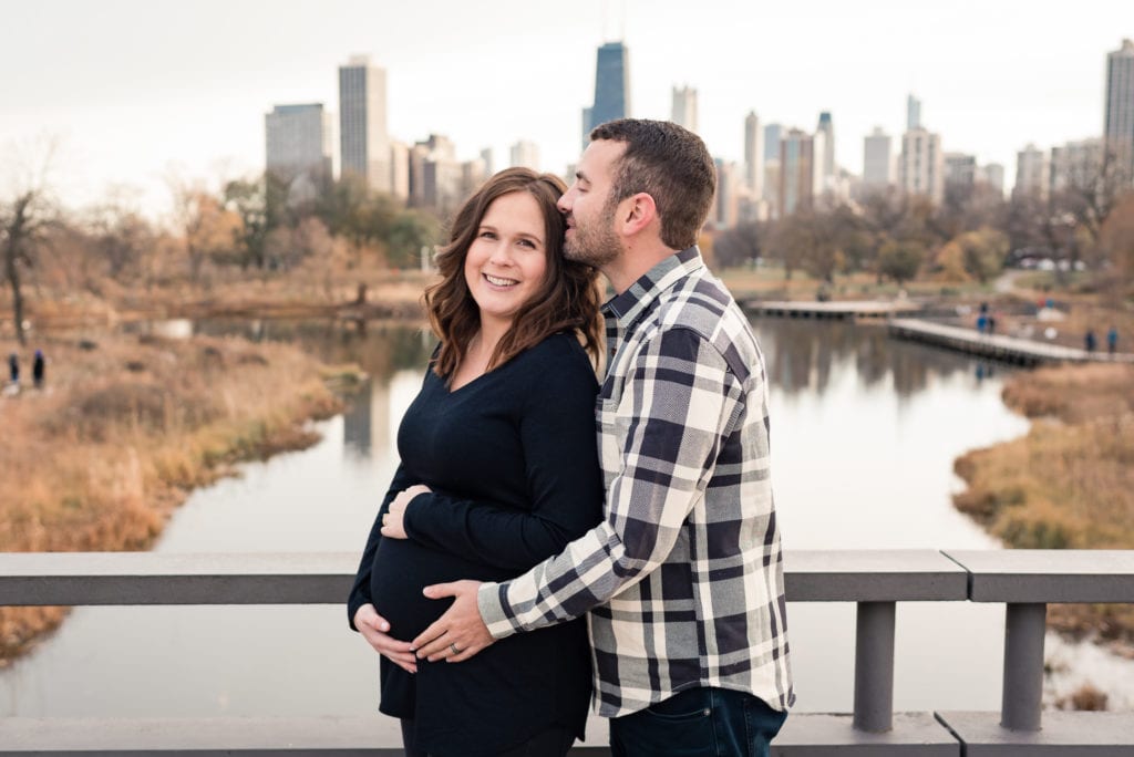 Husband kissing wife on temple for maternity pictures with chicago skyline in the background at Lincoln park nature boardwalk