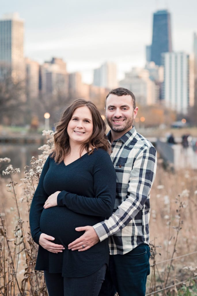 husband and wife smiling at camera in Lincoln park nature boardwalk with chicago skyline in the background for maternity pictures