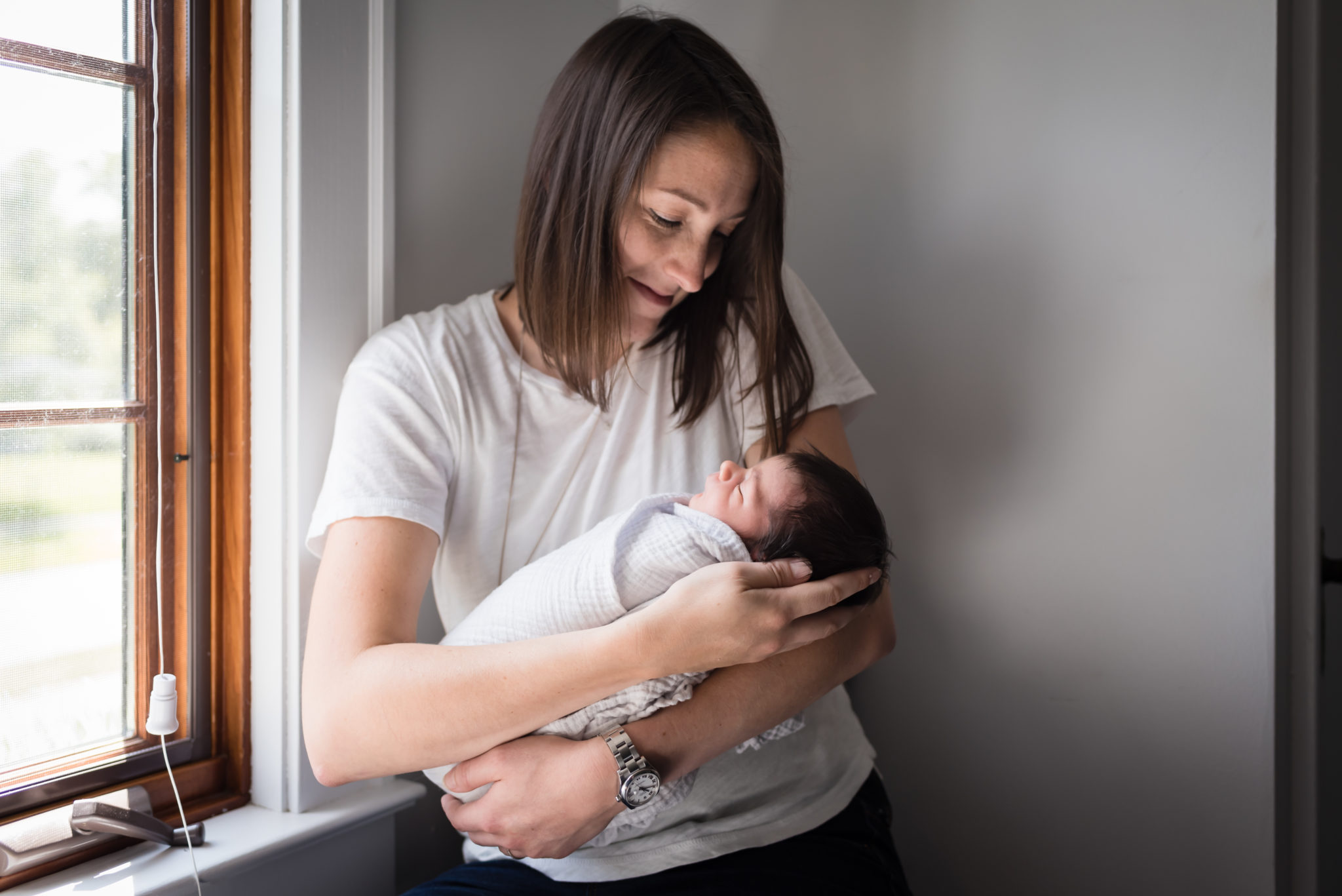 Mom with shoulder lengthy brown hair holding newborn in white wrap by a window